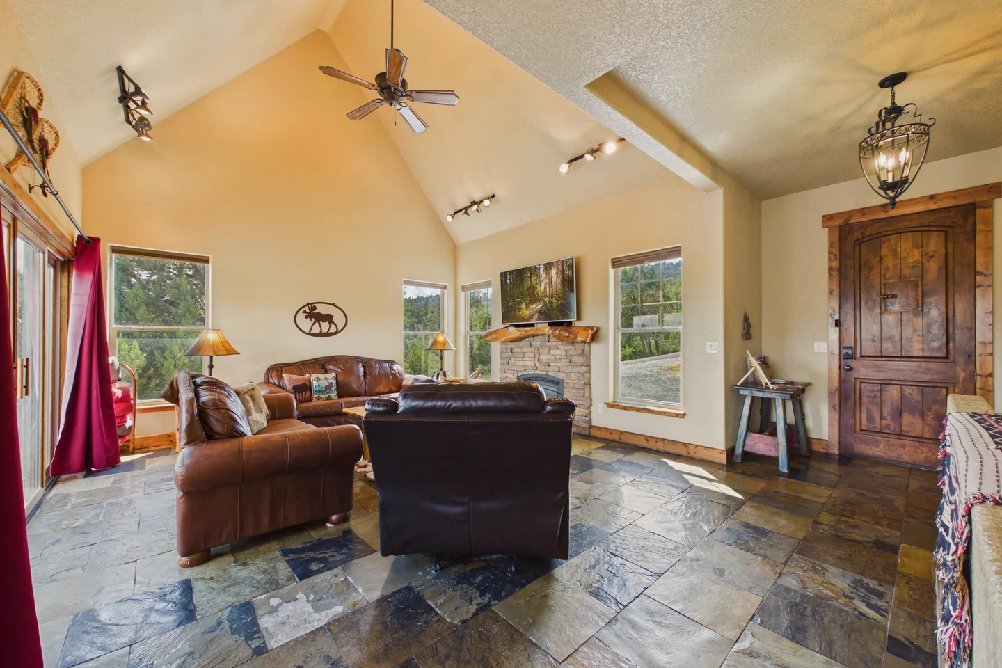 Living room with vaulted ceilings, leather seating, a stone-faced fireplace with a mounted TV, and slate-style tile flooring.