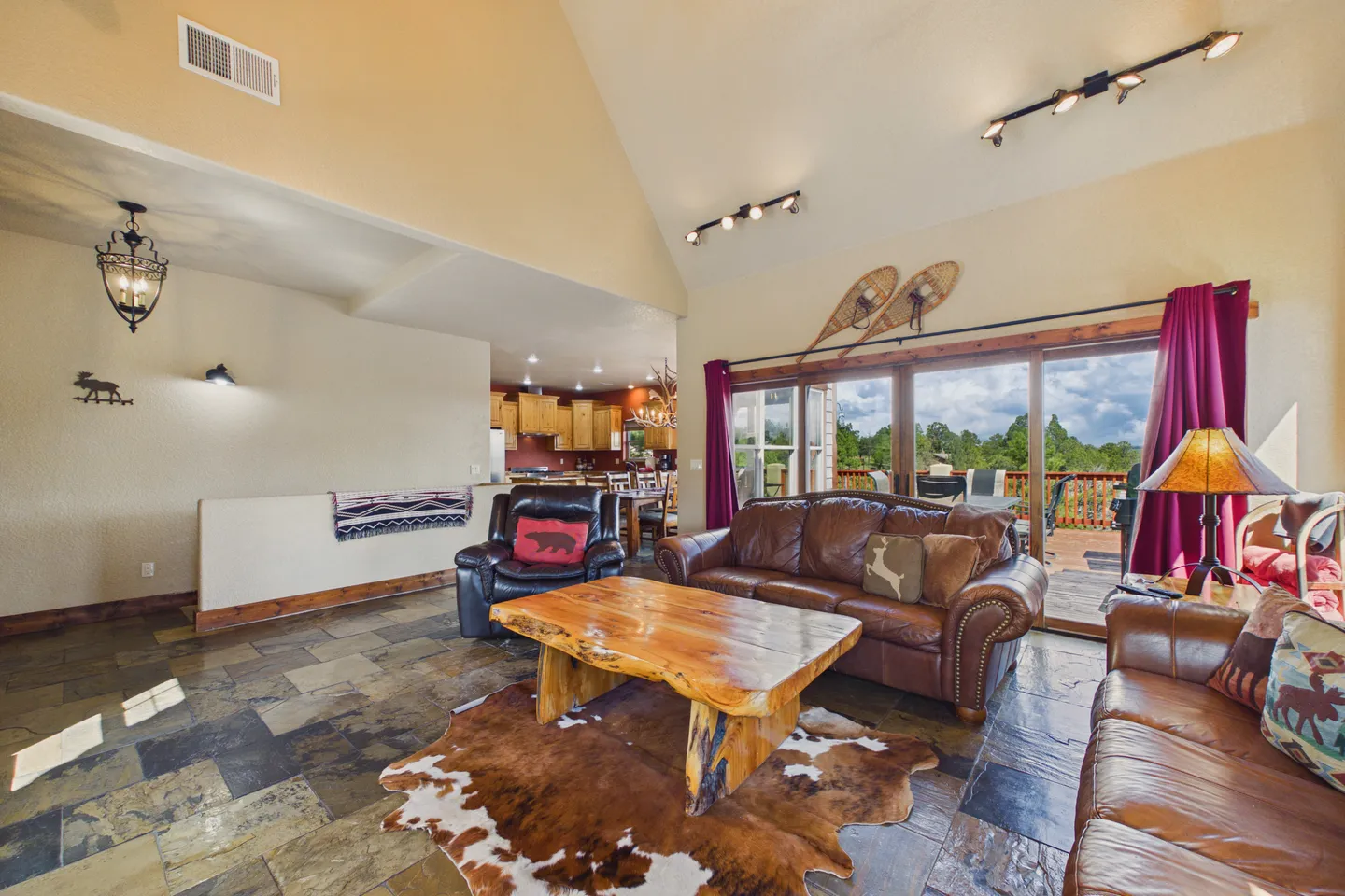 Living room with vaulted ceiling, brown leather sofas, a live-edge wood coffee table, tiled floors, and sliding glass doors opening to an outdoor deck.