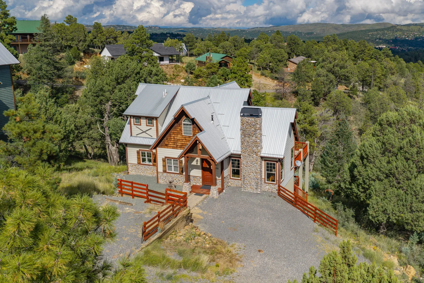 Aerial view of a two-story home with a metal roof, stone chimney, and wood fencing set in a hillside landscape with pine trees.