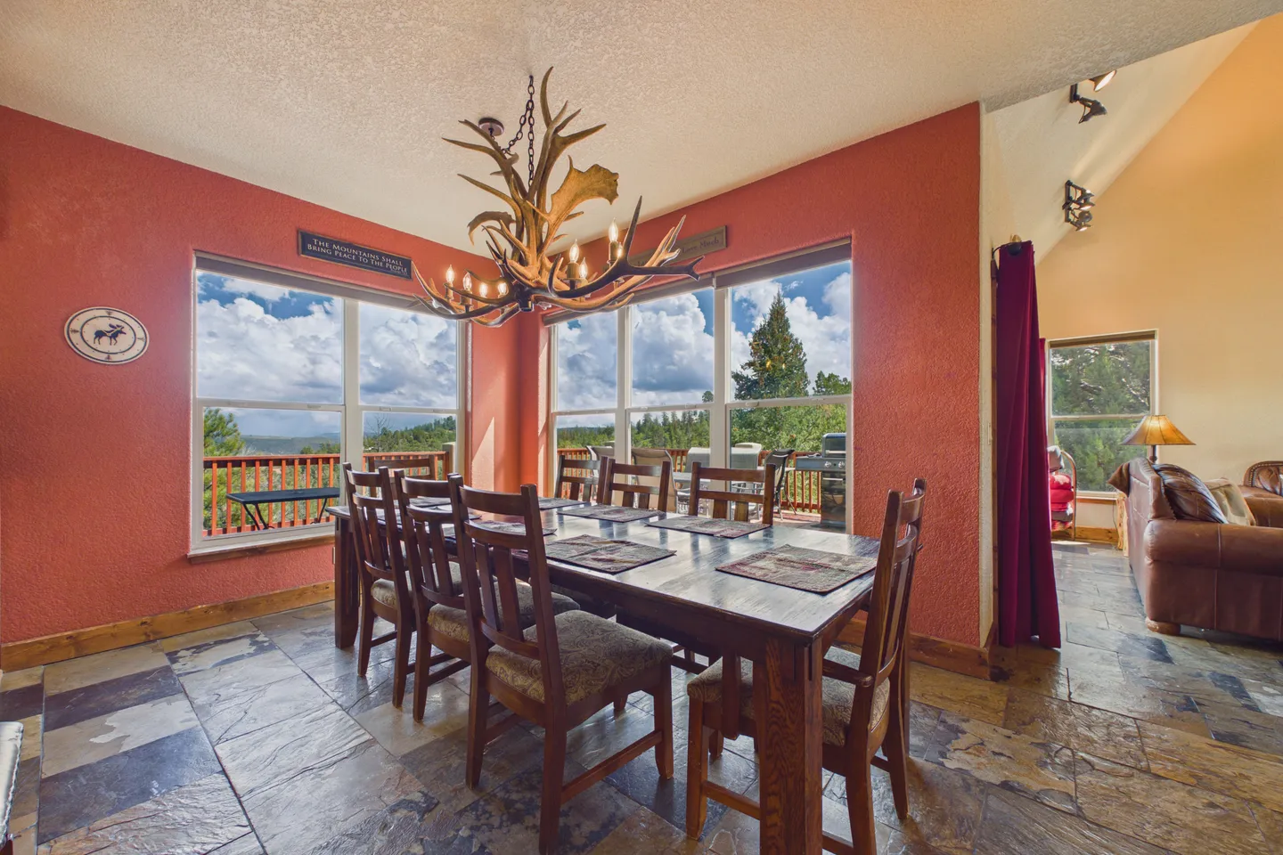 Dining room with a rustic antler chandelier, wood table and chairs, stone tile flooring, and large windows looking outside to a deck and trees.