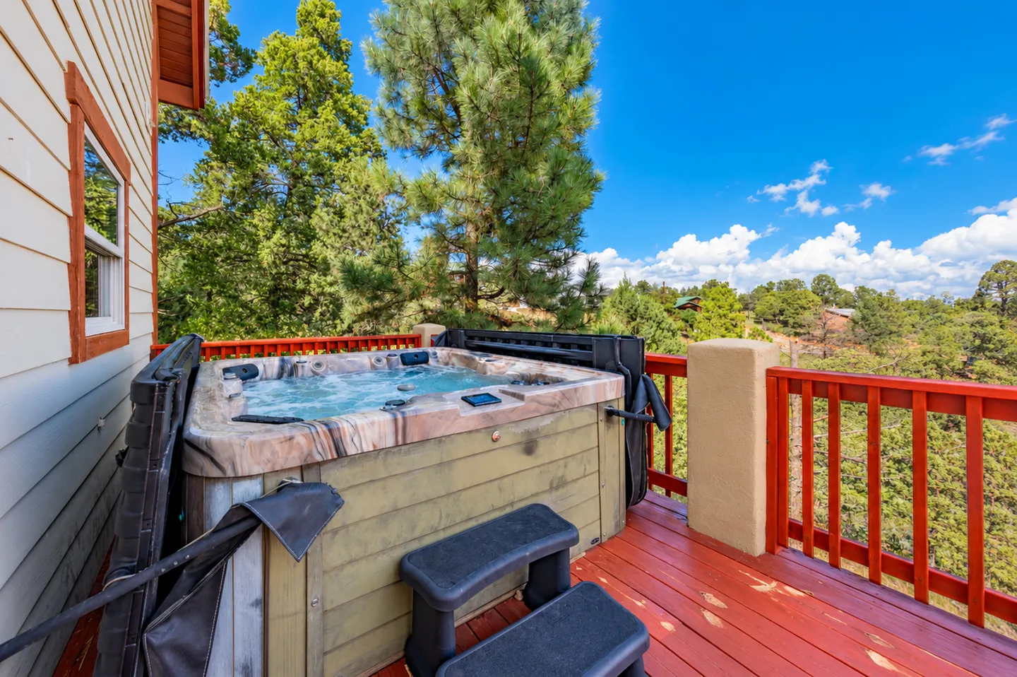 Outdoor hot tub on an elevated wood deck overlooking pine trees under a blue sky.