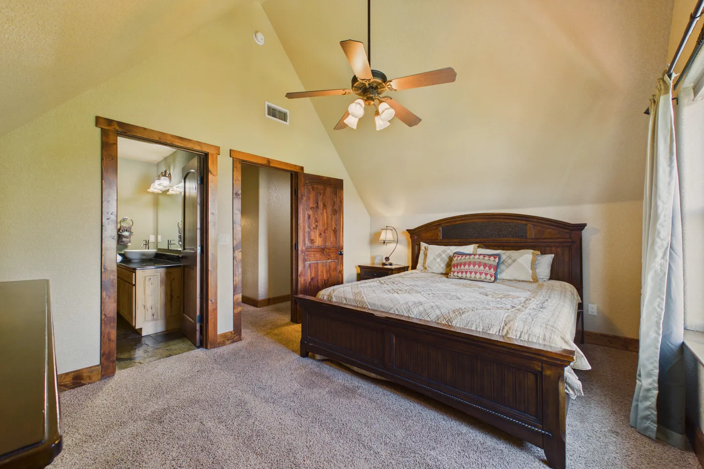 Bedroom with vaulted ceiling, ceiling fan, wooden bed frame, carpeted floor, and an open doorway leading to an en-suite bathroom.