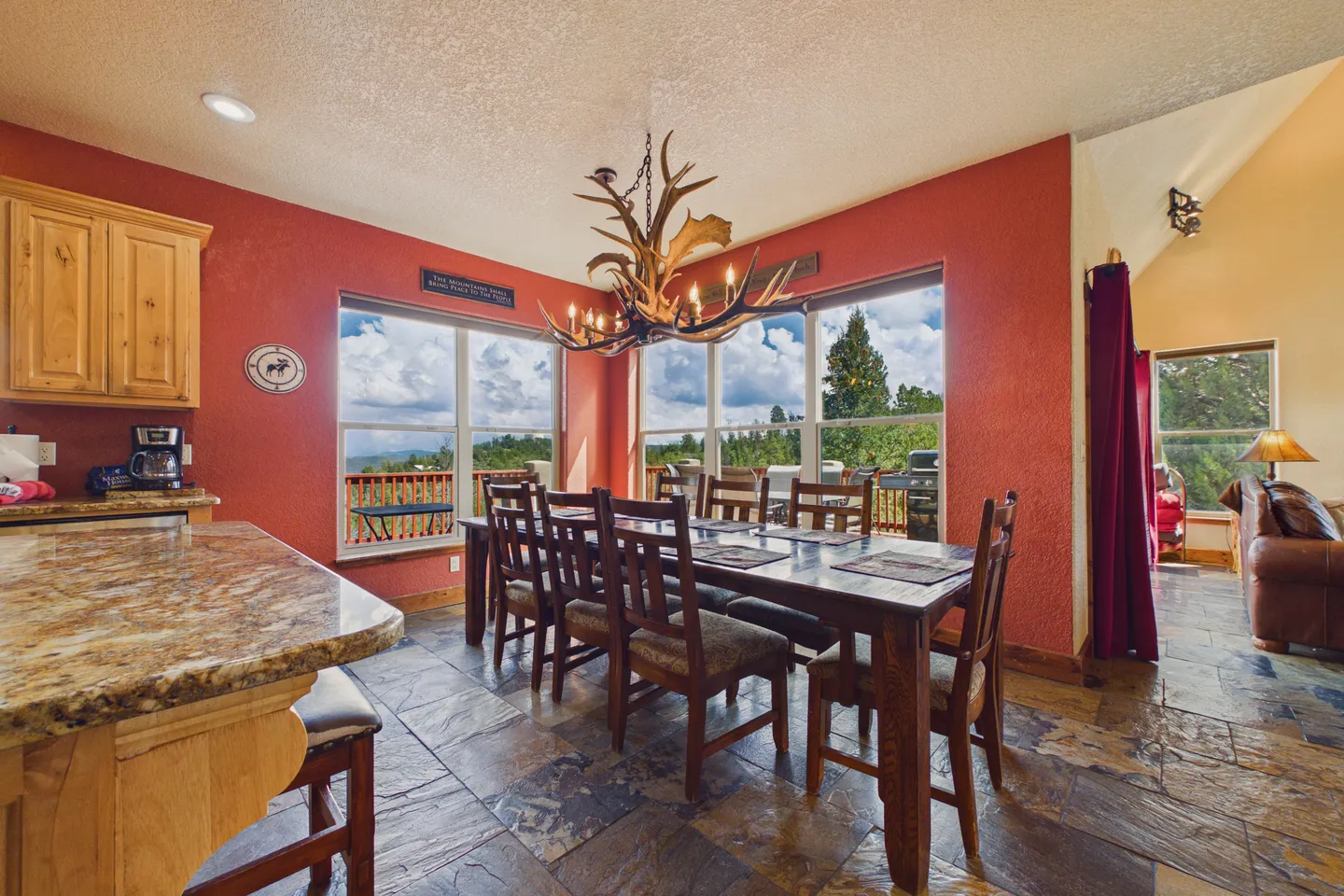 Dining area with a large wooden table, antler chandelier, and red accent walls next to large windows.