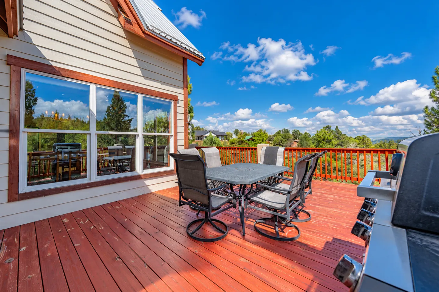 Outdoor wooden deck with a dining table, chairs, barbecue grill, and view of surrounding trees under a blue sky.