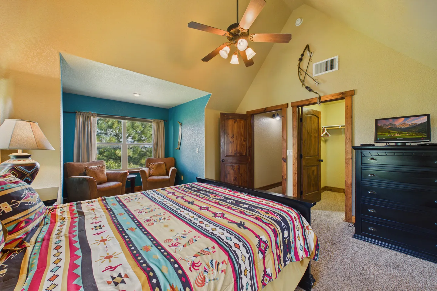 Bedroom with vaulted ceiling, patterned bedding, two armchairs in a blue alcove, ceiling fan, and black dresser with a television.