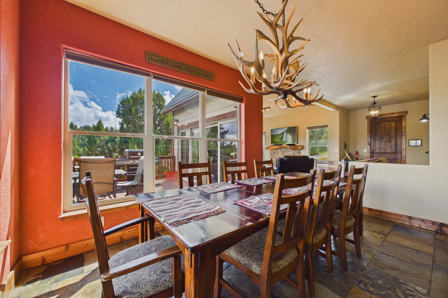 Dining room with a long wooden table, antler chandelier, terracotta-colored accent wall, and large windows looking onto an outdoor deck.