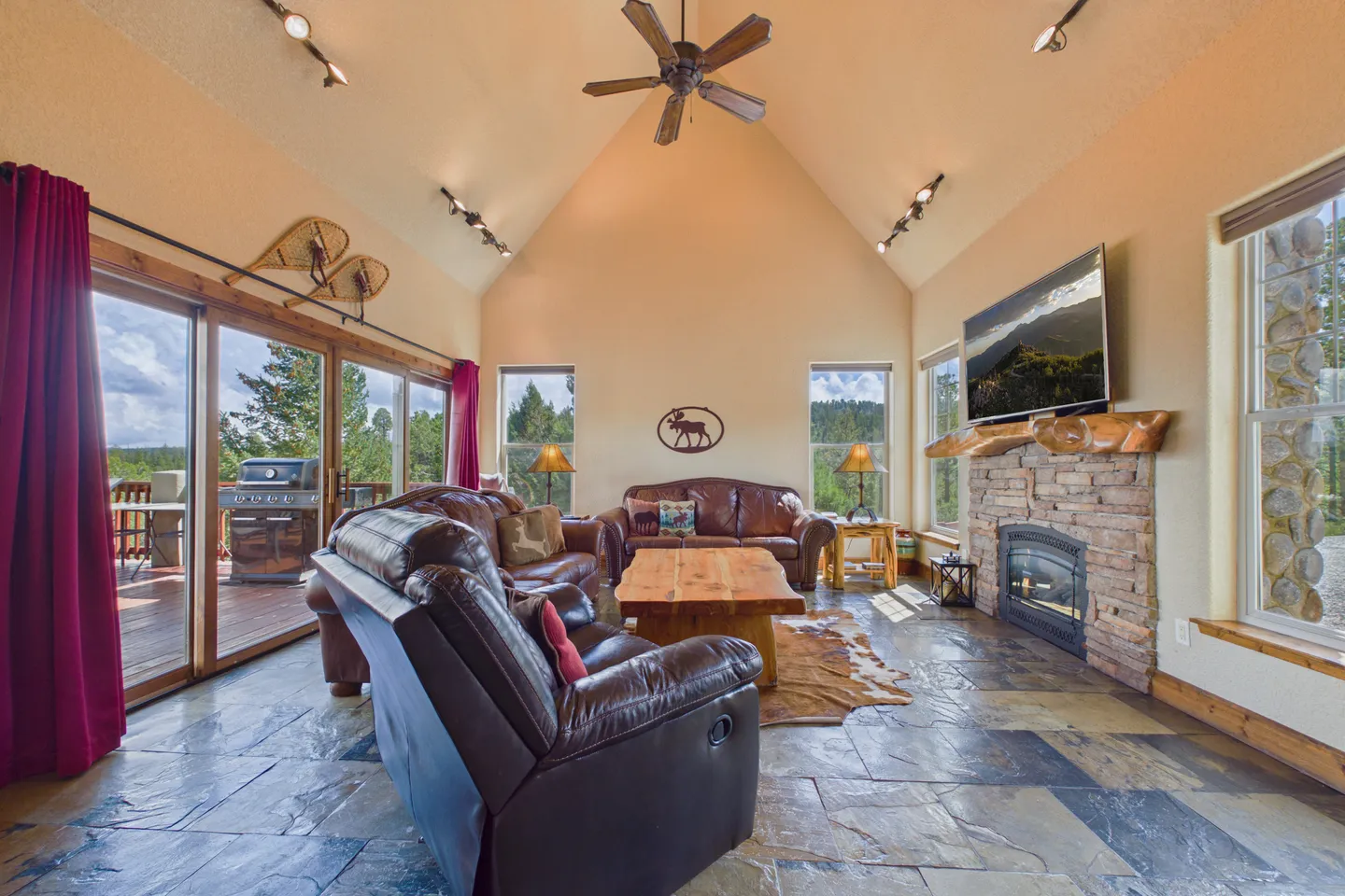 Living room with high vaulted ceiling, stone fireplace, leather seating, and sliding glass doors leading to an outdoor deck.