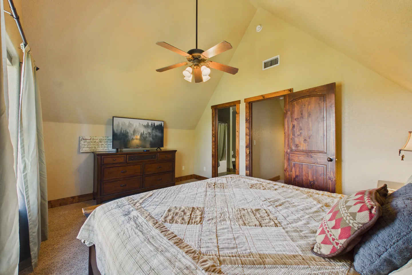 Bedroom with vaulted ceiling, ceiling fan, bed with patchwork quilt, TV on a wooden dresser, and an open wooden door leading to an en-suite bathroom.