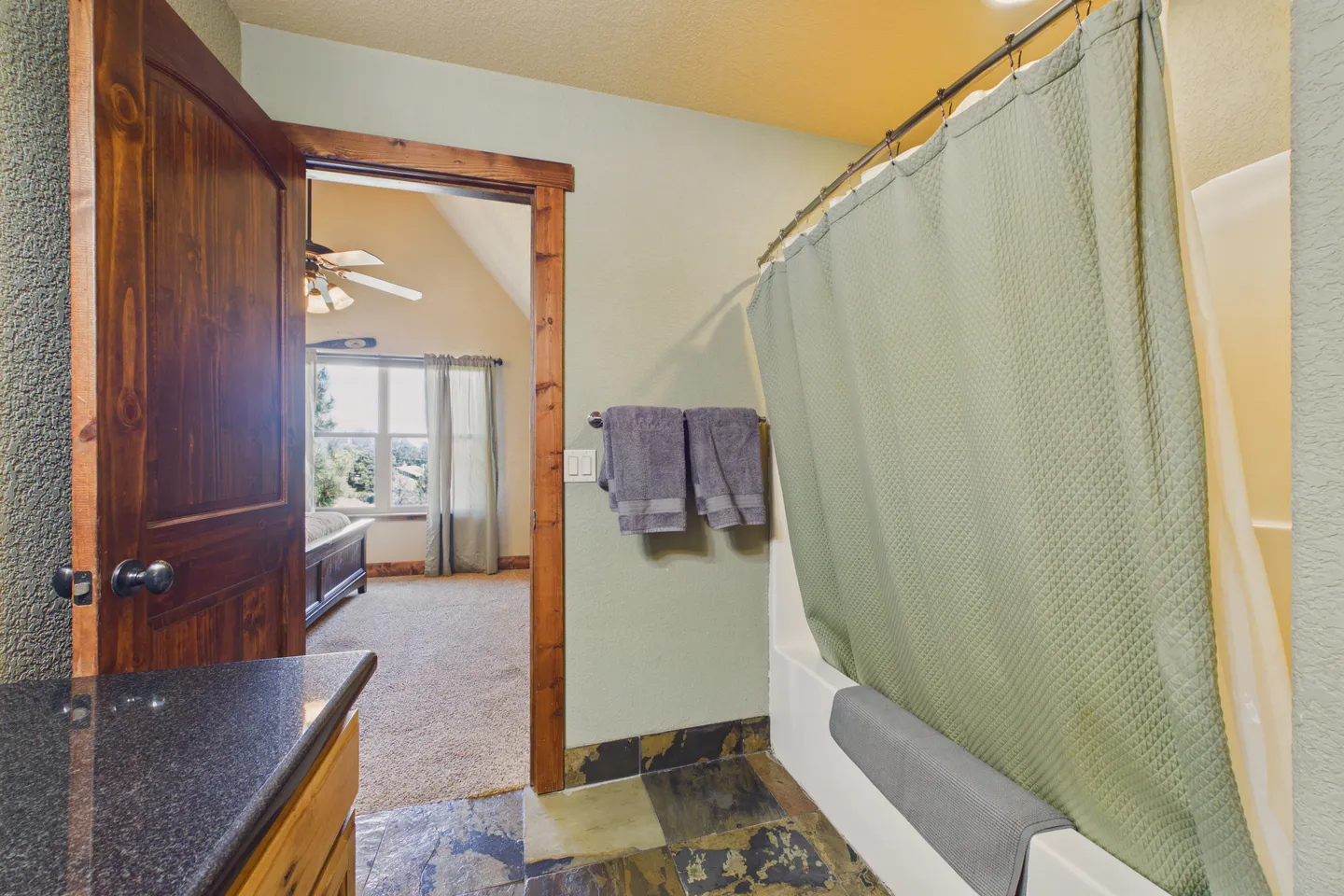 Bathroom with slate tile flooring, a bathtub with a green curtain, and an open doorway looking into a carpeted bedroom.