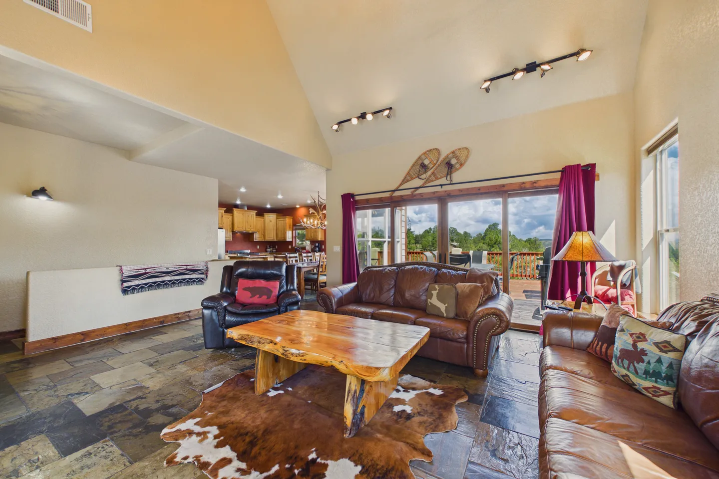 Living room with vaulted ceiling, brown leather couches, rustic wood coffee table, tiled floor, and sliding doors to an outdoor deck.