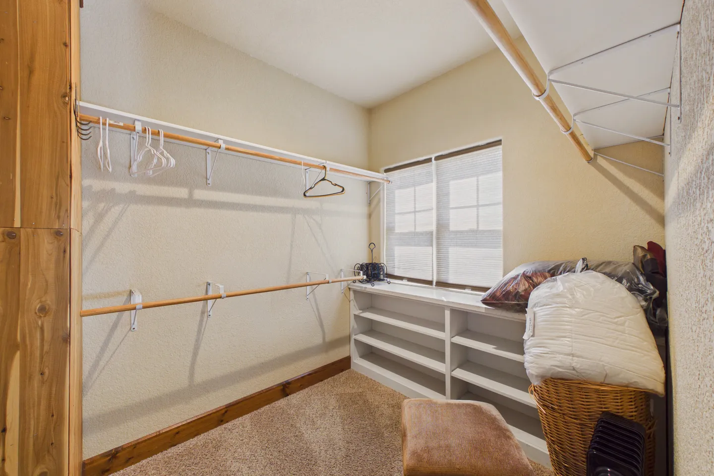 A walk-in closet with carpet, two tiers of clothing rods, built-in low shelves, and a window.