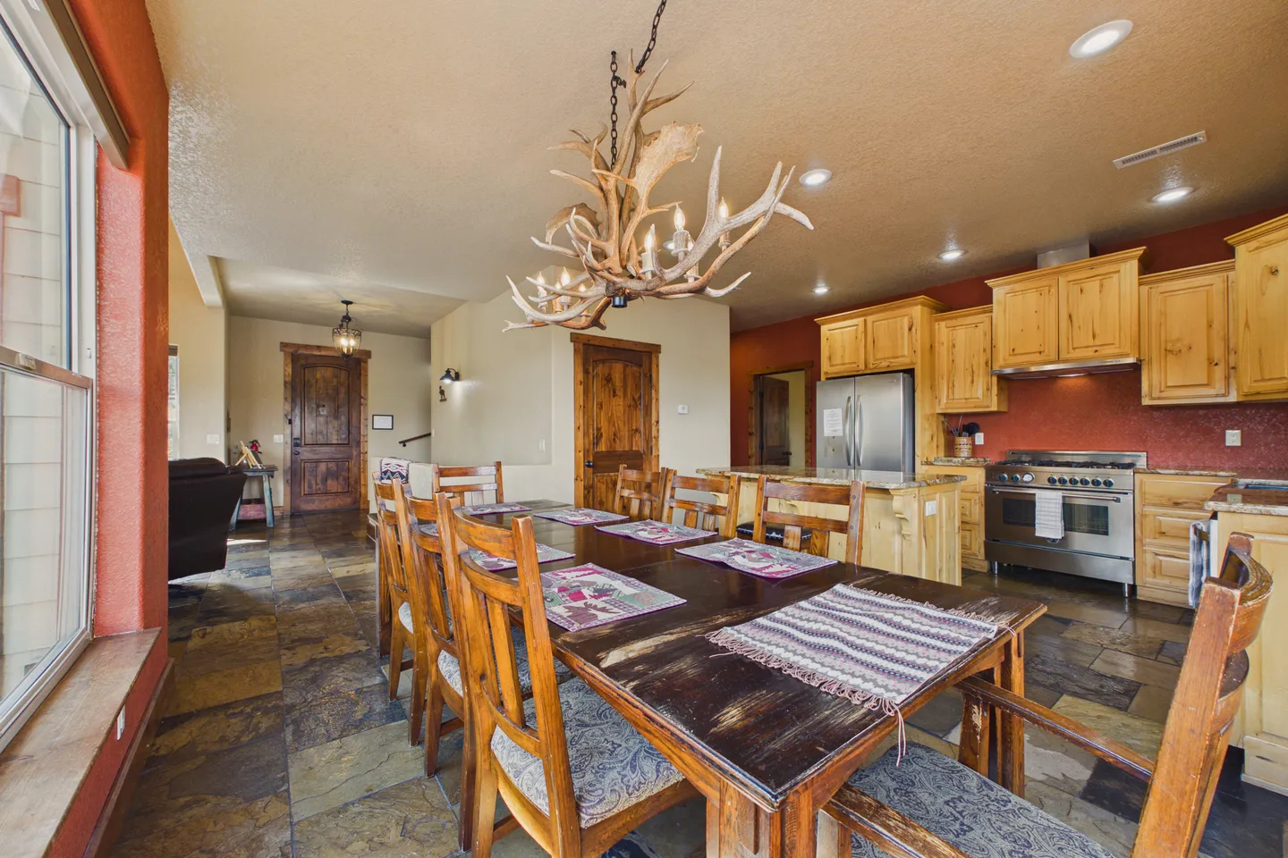Dining area with a large wooden table, antler chandelier, tiled floors, and an adjacent kitchen with light wood cabinets.