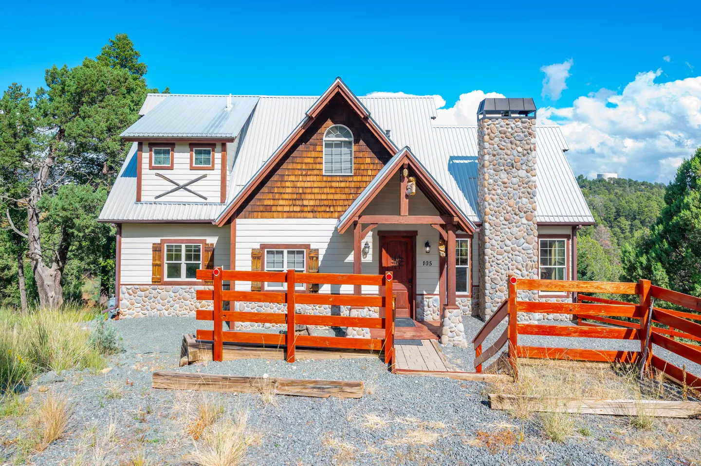 Exterior front view of a two-story home with a metal roof, stone chimney, wood siding, and wooden railings under a blue sky.