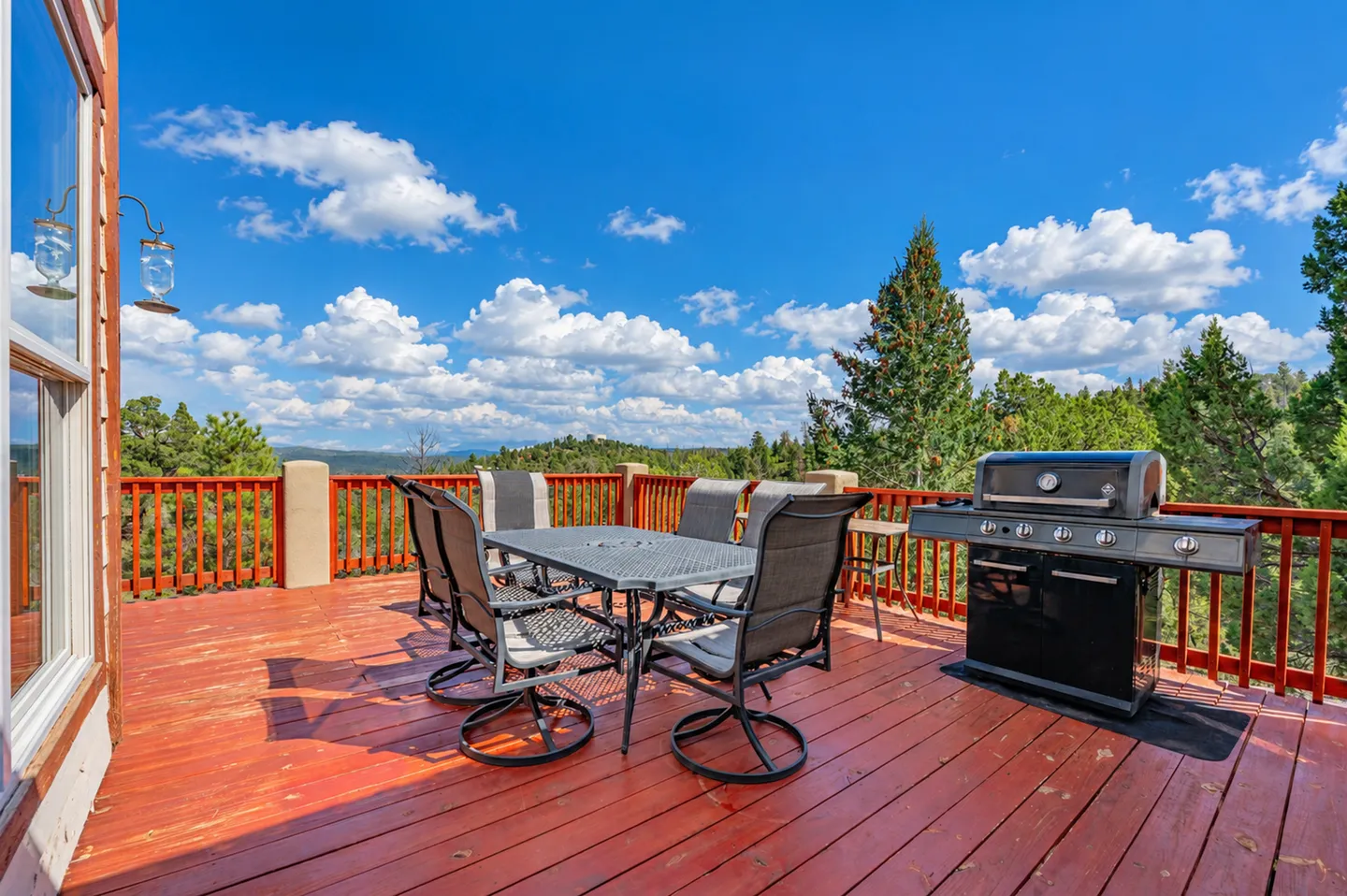 Outdoor wooden deck featuring a dining table with chairs, a gas grill, and views of surrounding evergreen trees and mountains under a blue sky.