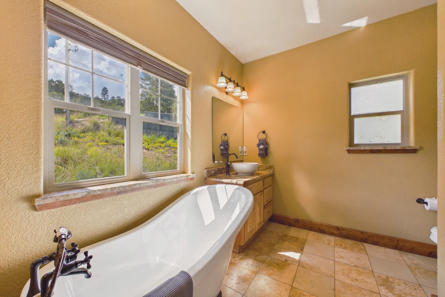 Bathroom with a white freestanding soaking tub, tiled floor, and a vanity beneath windows overlooking a hillside.