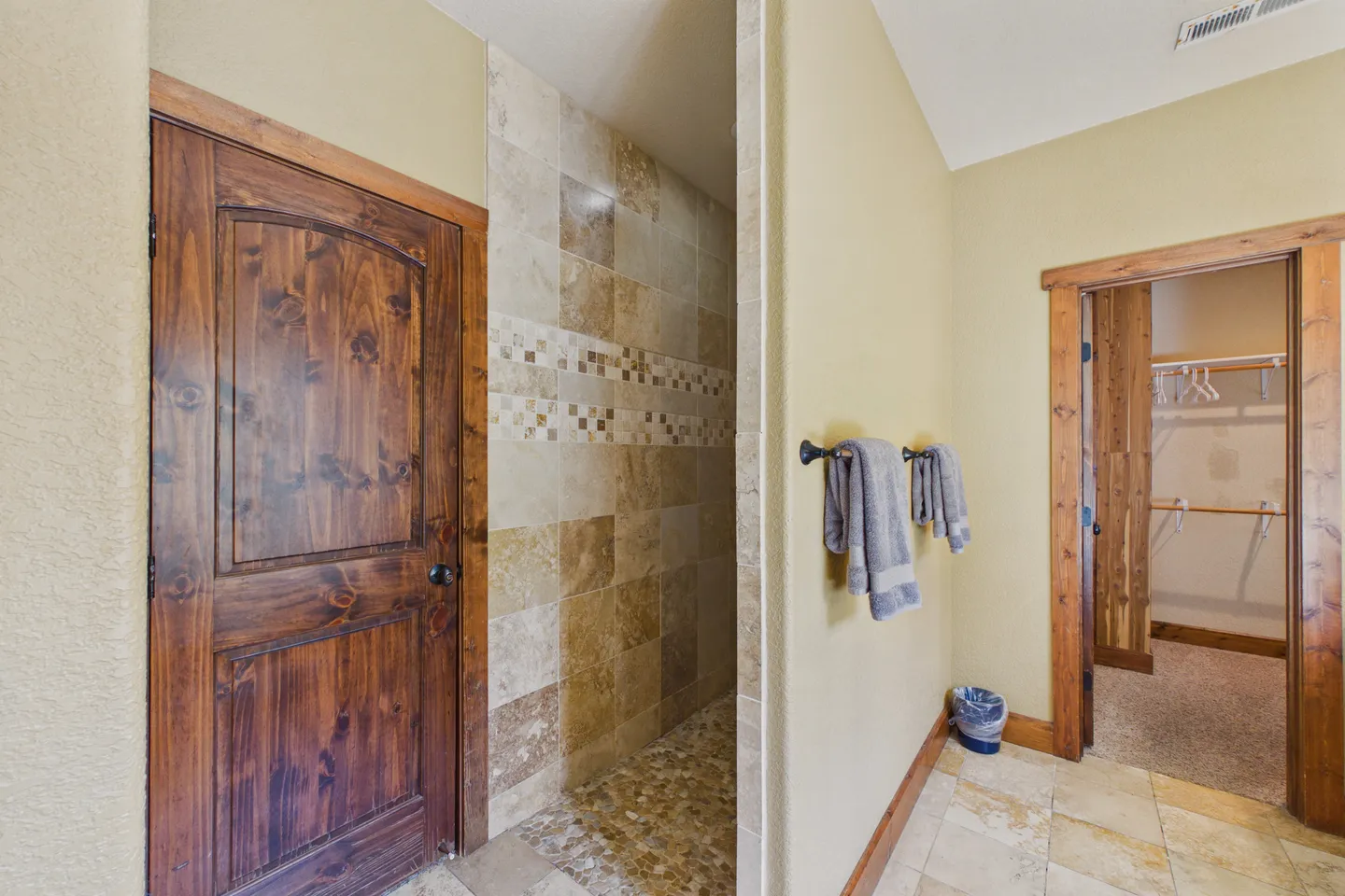 Bathroom showing a tiled walk-in shower entrance, wooden doors, towel hooks with towels, and an open doorway leading to a walk-in closet.