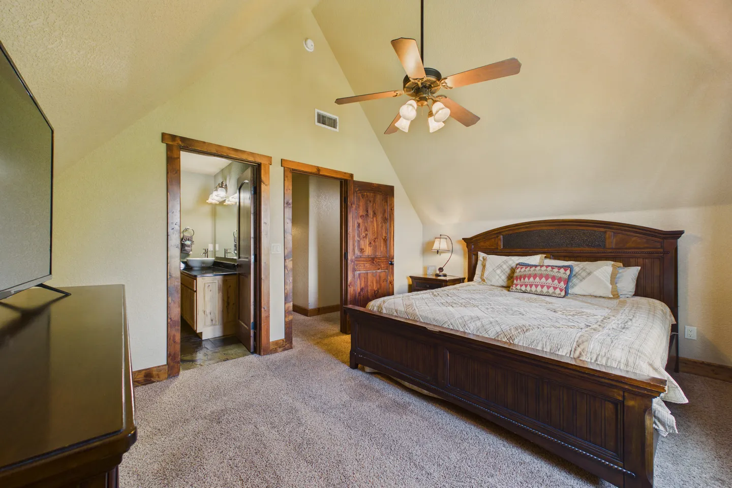 Bedroom with vaulted ceiling, large wood bed frame, ceiling fan, carpet, and doorway leading into an en suite bathroom.