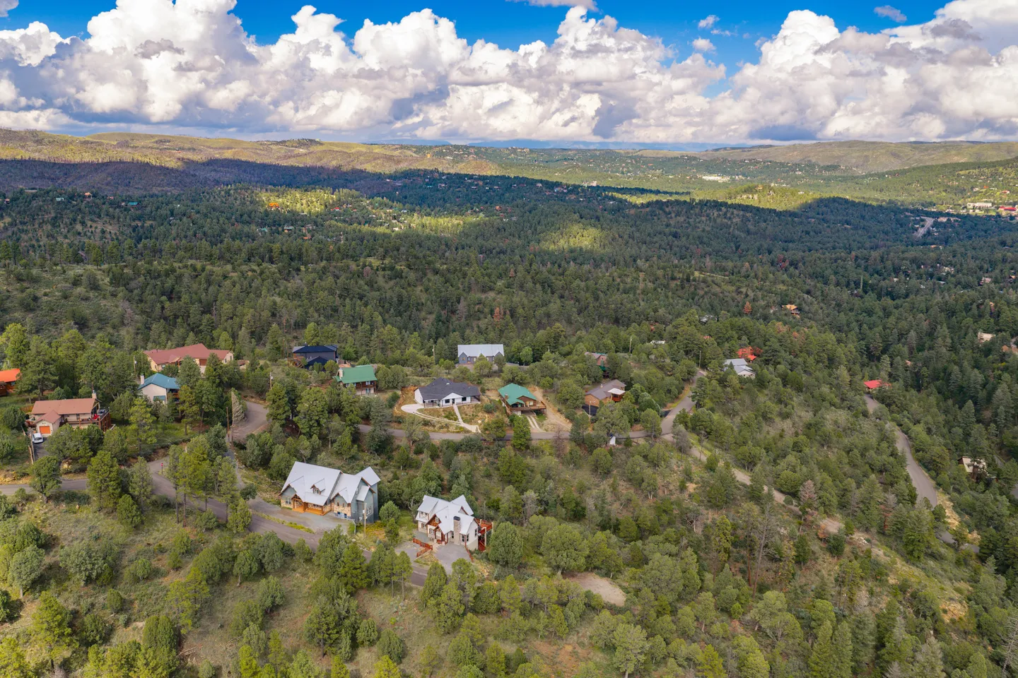 Aerial view of homes nestled on a densely forested hillside under a blue sky with fluffy white clouds.