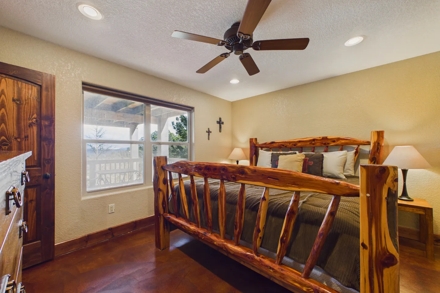 Bedroom featuring a natural wood log-style bed frame, matching nightstands with lamps, a ceiling fan, and a window with views of an outdoor deck.