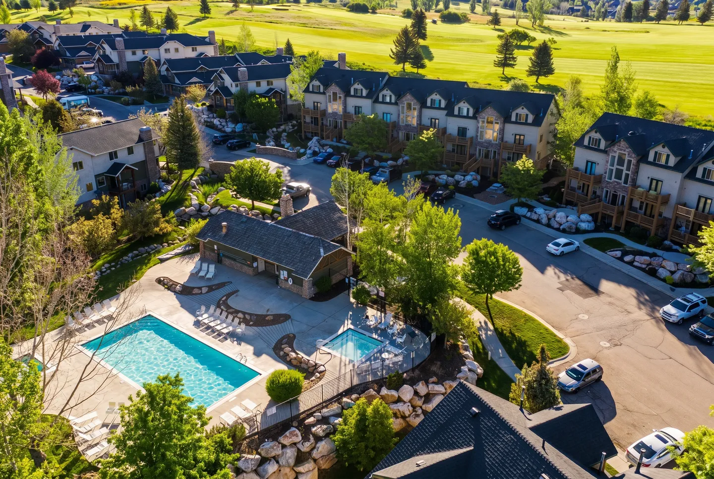 Aerial view of a residential community featuring a swimming pool, hot tub, club building, and multi-story townhomes bordered by green fields.