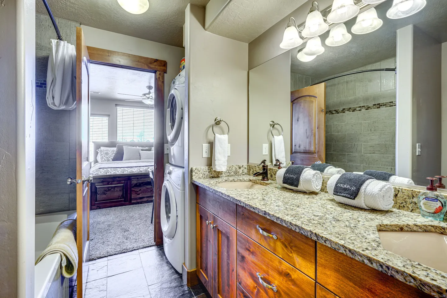 Bathroom with double sink vanity, stacked washer and dryer, tiled shower-tub combo, and an open doorway leading to a bedroom.