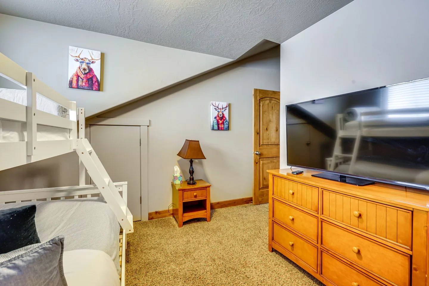 Bedroom featuring a white bunk bed, carpeted flooring, a wooden dresser with a television, and sloped ceiling sections.