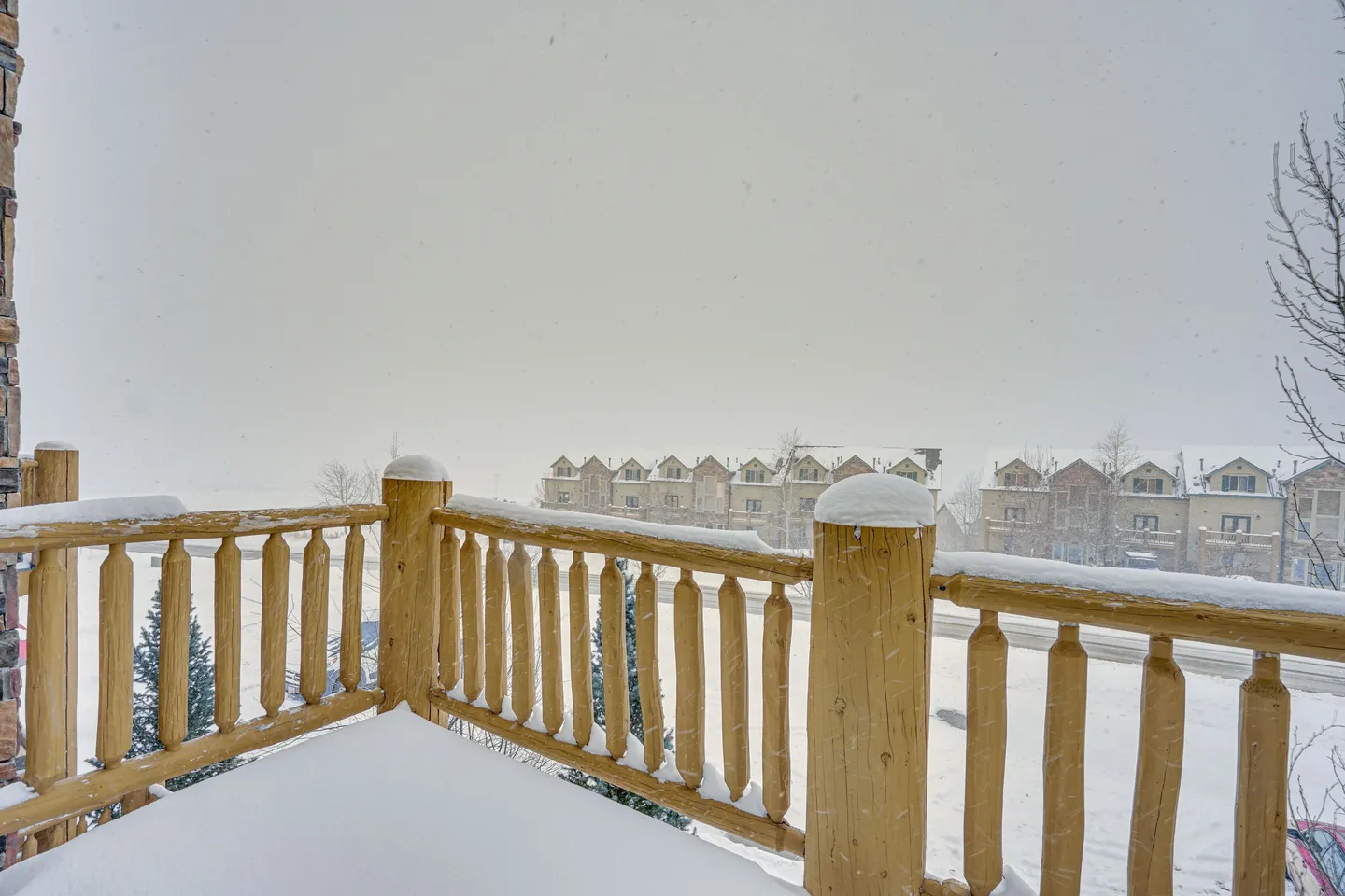 Snow-covered wooden balcony with a log railing looking out over townhouses during a snowfall.