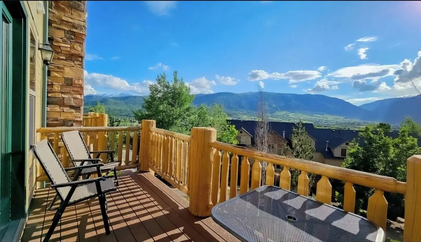A wooden balcony with rustic log railings, outdoor chairs, and a table overlooking a vast valley and mountains under a blue sky.