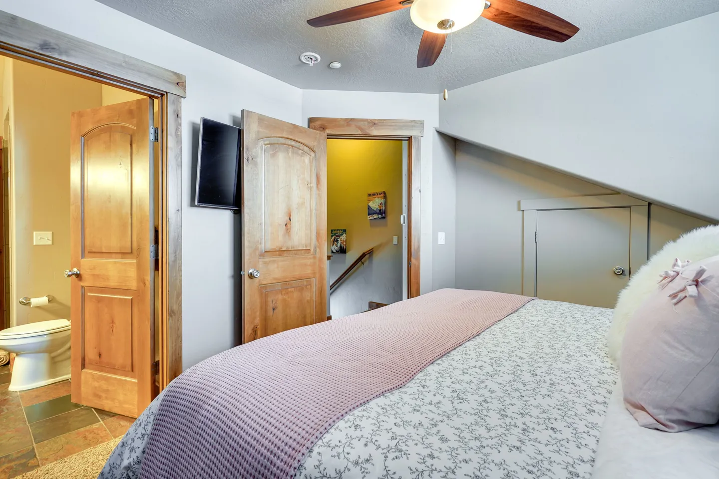 Bedroom with angled ceiling, ceiling fan, mounted television, and views into an en-suite bathroom and hallway.