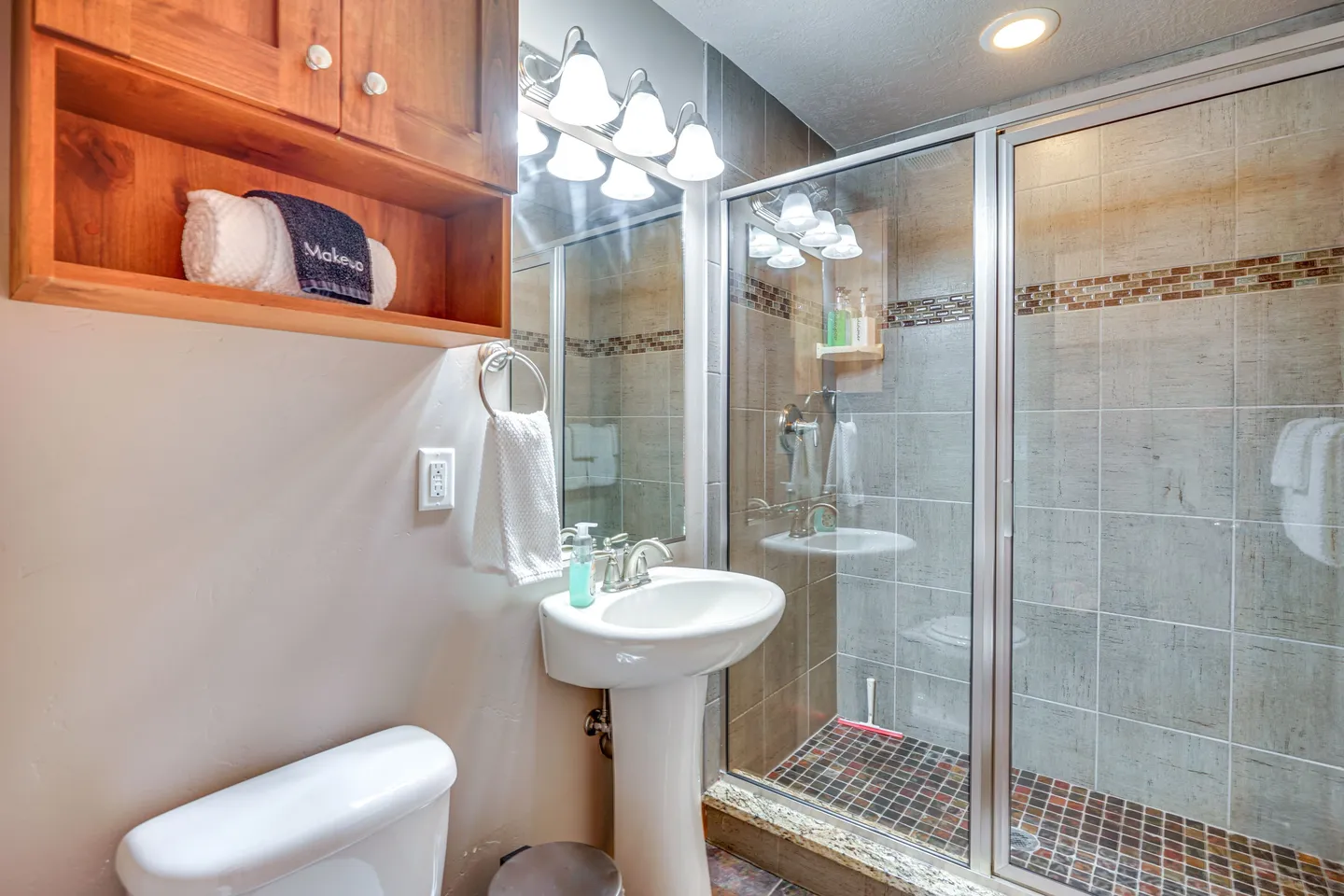 Bathroom with a pedestal sink, framed mirror with overhead vanity lighting, toilet, and glass-enclosed tiled shower.