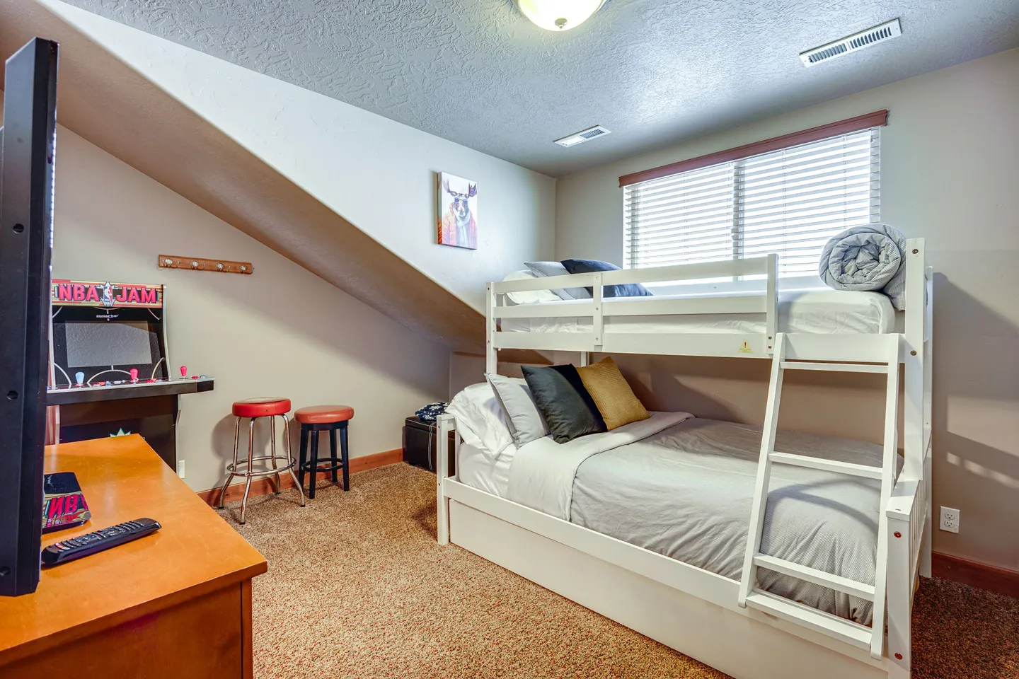 Bedroom with a white bunk bed, carpet flooring, a window with blinds, and an arcade cabinet with two stools under a sloped wall.