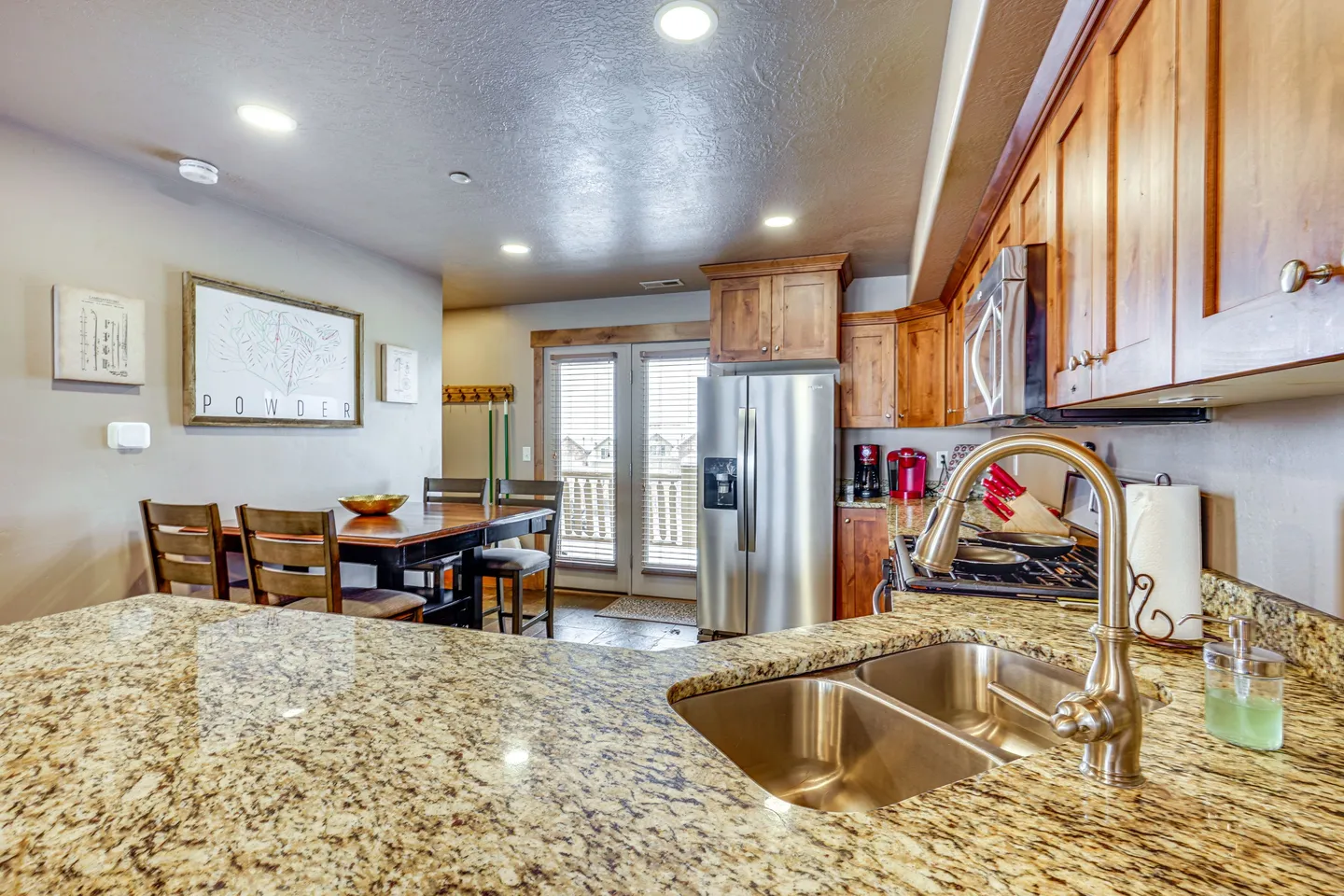 Kitchen counter and sink in the foreground looking toward a dining table and stainless steel refrigerator.