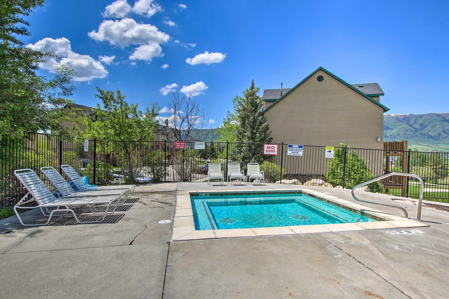 Outdoor square hot tub set in a concrete patio with lounge chairs and residential buildings behind a fence.