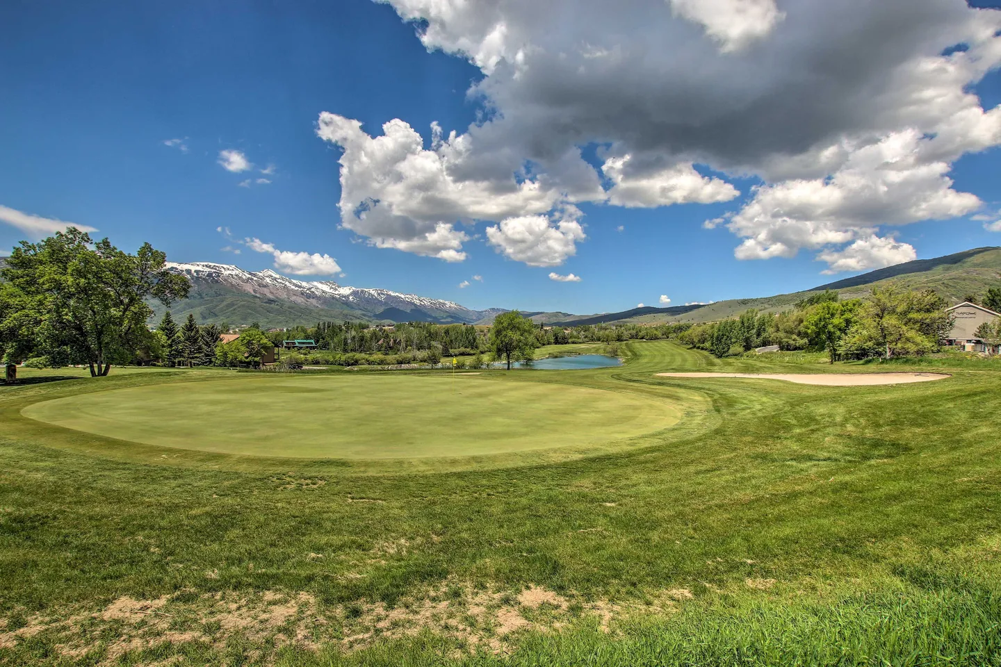 A wide view of a golf course green surrounded by rough, a sand trap, a small pond, and snow-capped mountains under a partly cloudy blue sky.