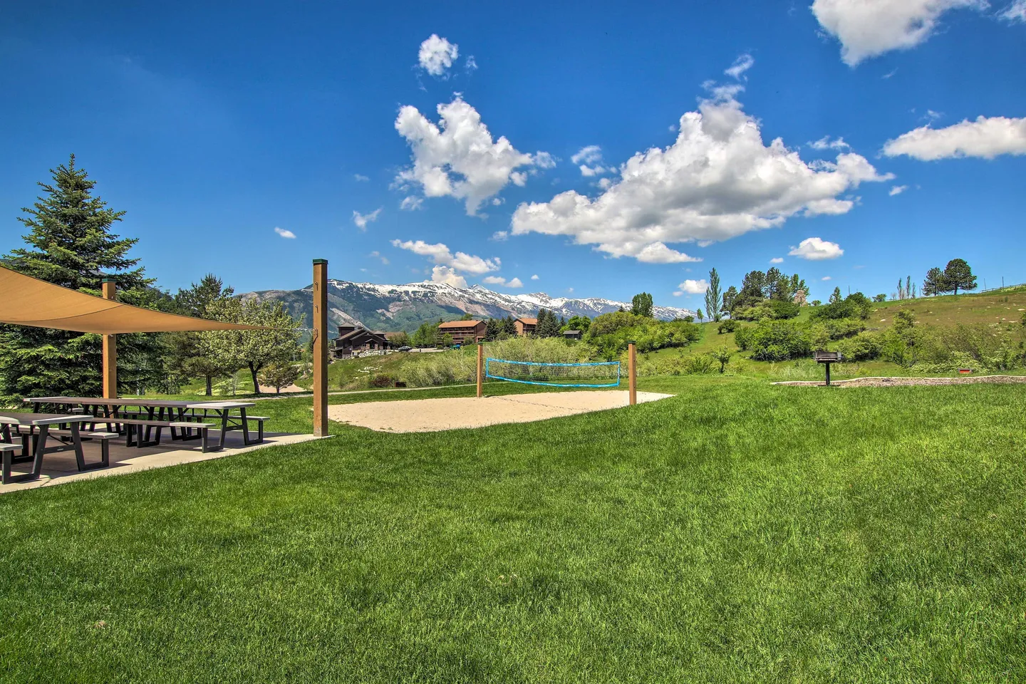 Outdoor sand volleyball court and shaded picnic tables on a grassy lawn with mountain views in the background.