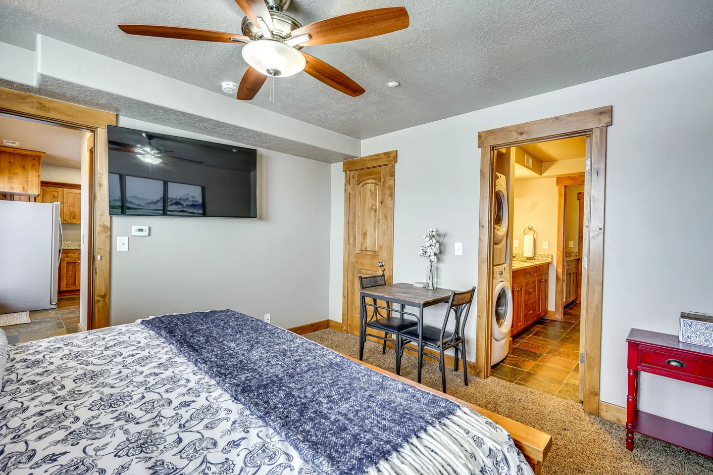 Bedroom featuring a bed with patterned bedding, a ceiling fan, wall-mounted television, small dining table, and doorway leading to an en-suite bathroom with laundry.
