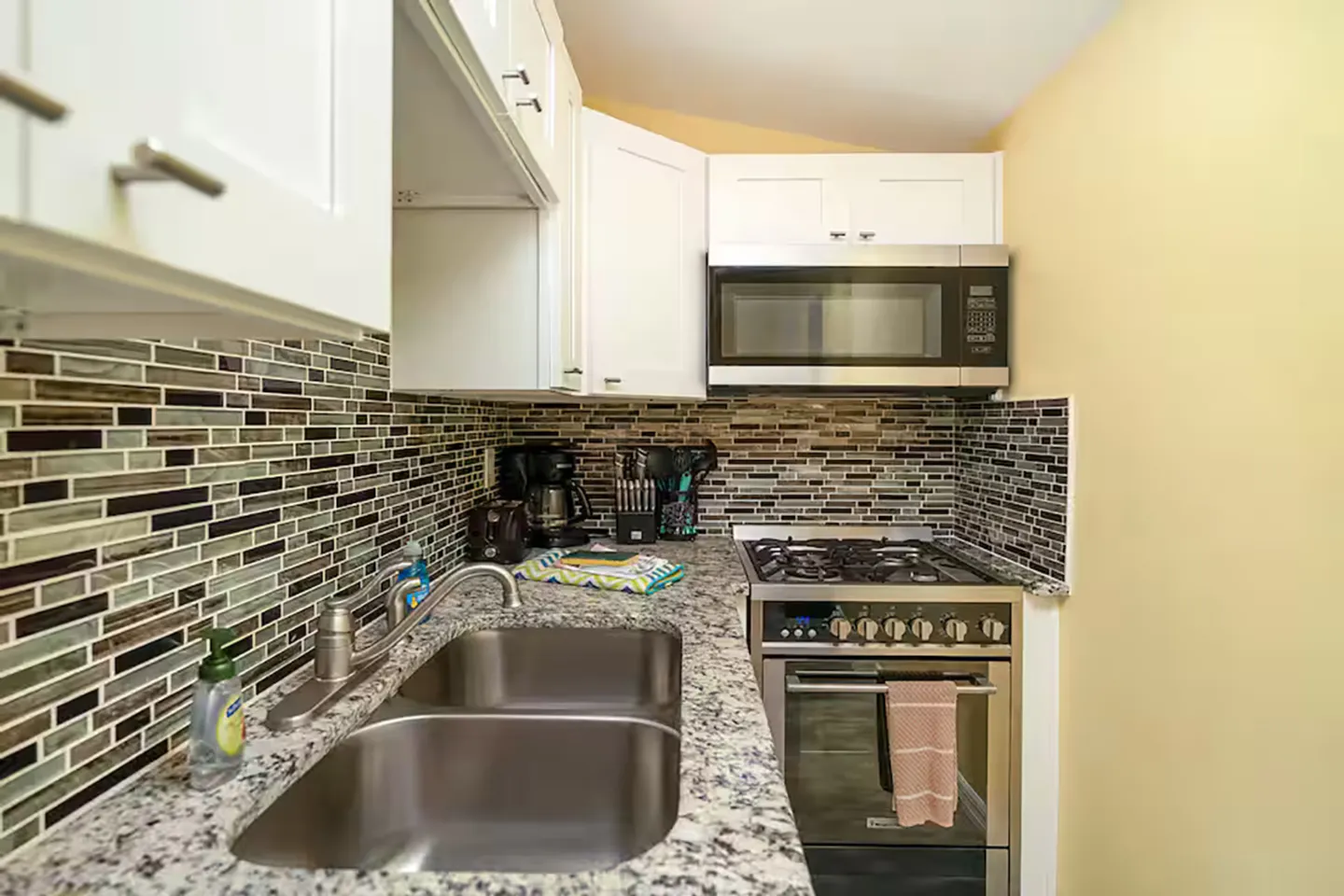 A compact kitchen featuring a double stainless steel sink, speckled granite-look countertops, tiled backsplash, white cabinetry, and stainless steel stove and microwave.