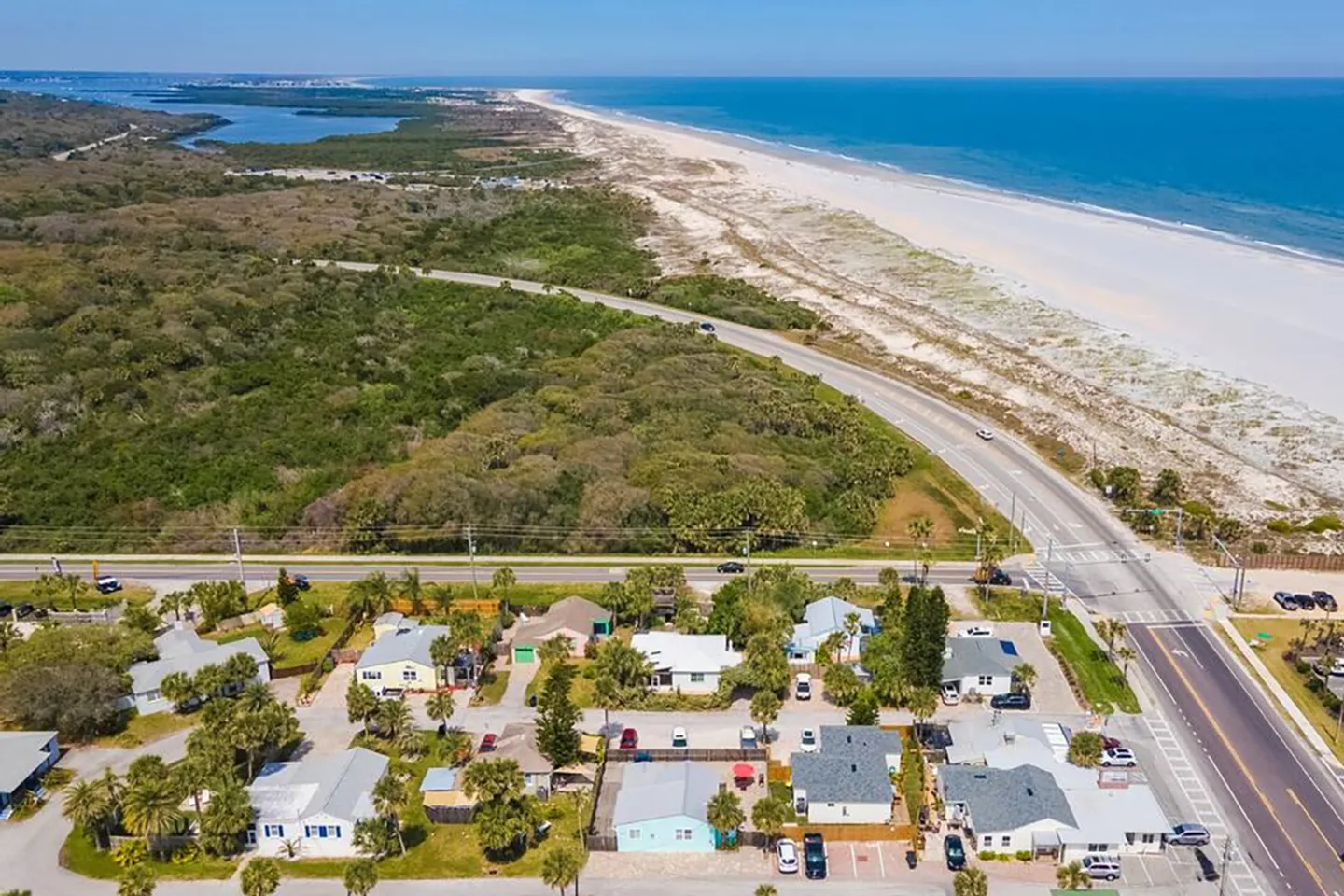 Aerial view of a coastal neighborhood situated across a roadway from sand dunes and an ocean beach.