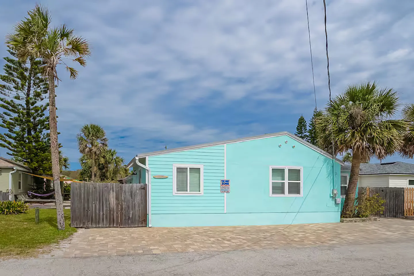 Exterior view of a light blue single-story home with a paved driveway, wooden fencing, and surrounding palm trees under a blue sky.