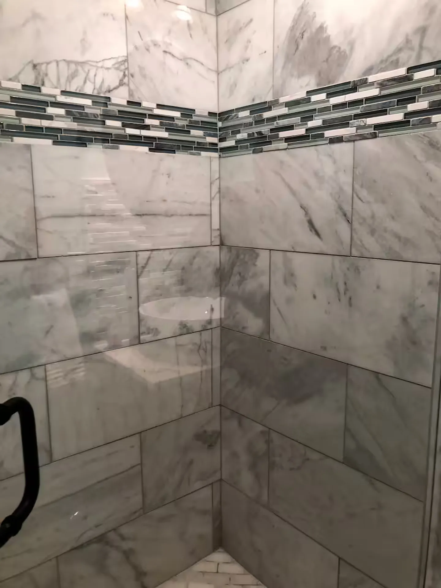 Close-up view of a tiled shower wall with marble-patterned tiles and a decorative glass mosaic accent band.