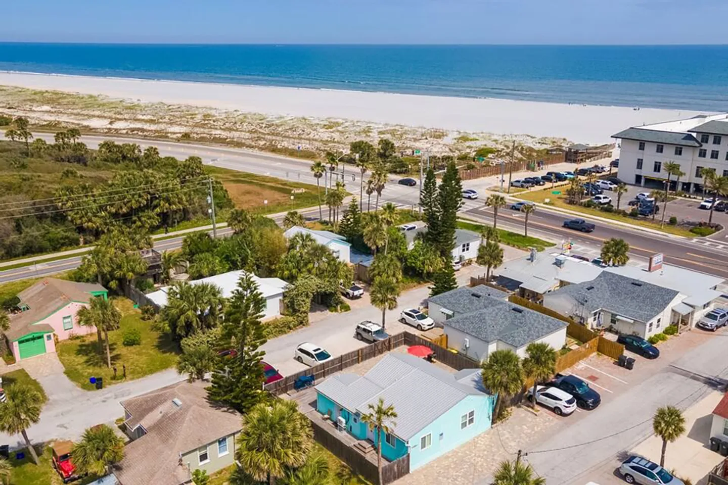 Aerial view of a coastal neighborhood with single-story homes across a road from a white sand beach and the ocean.