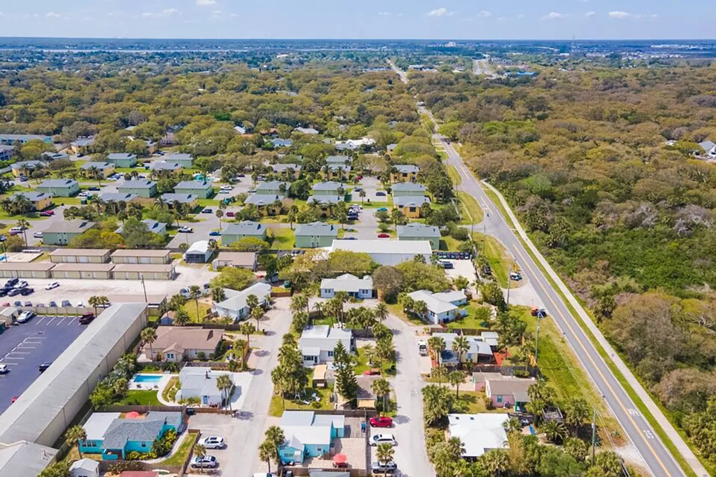 Aerial view of a coastal neighborhood with single-story homes, palm trees, and an adjacent multi-lane road running through a lush green area.