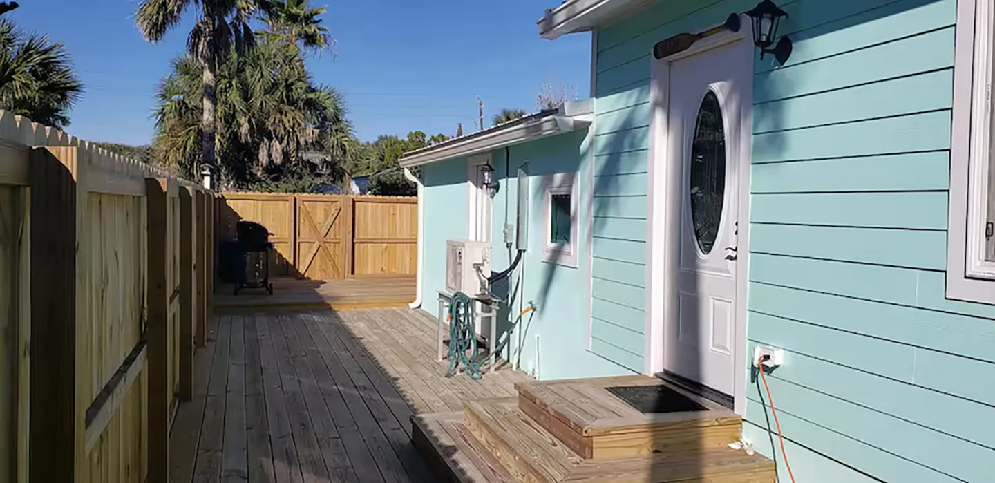 Exterior wooden deck alongside a light teal house enclosed by a tall wooden fence, with an entrance door and steps.