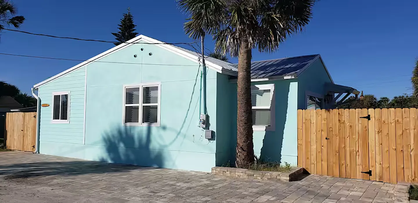 Exterior view of a light blue single-story home with a paved driveway, palm tree, and wooden privacy fence under a clear blue sky.