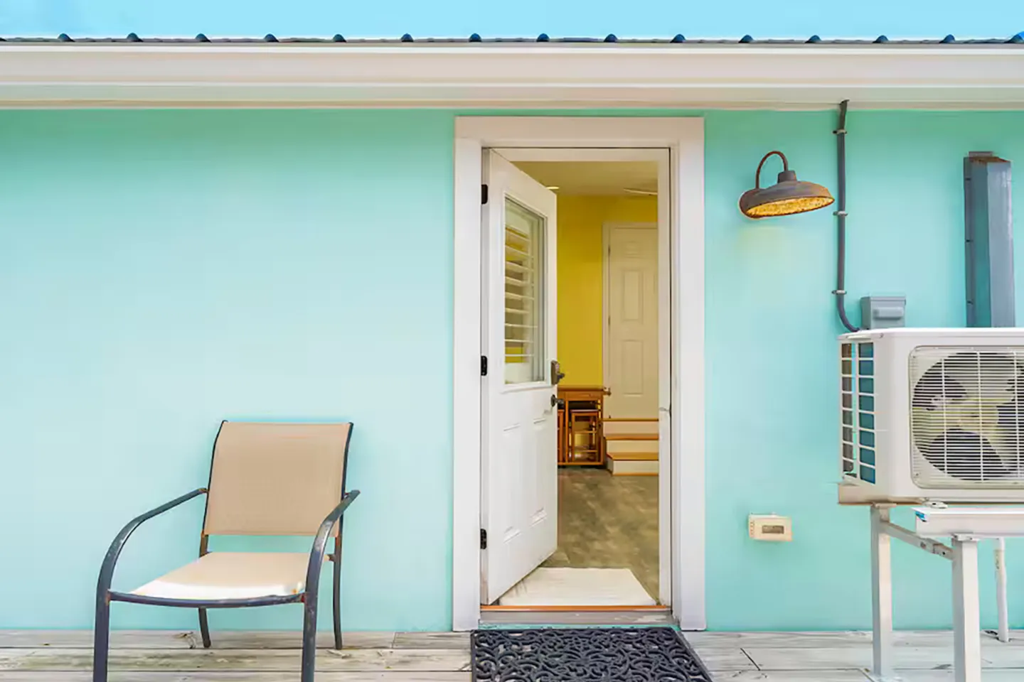 Exterior light-blue wall with an open door leading inside to a hallway, next to a patio chair and an exterior AC condenser unit.