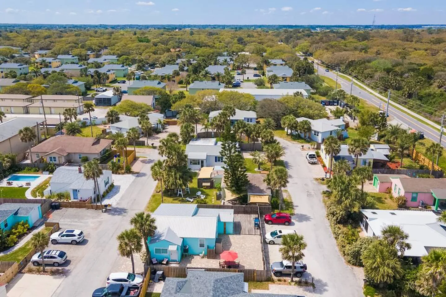 Aerial view of a coastal neighborhood with colorful one-story homes, palm trees, and parked cars under a clear sky.