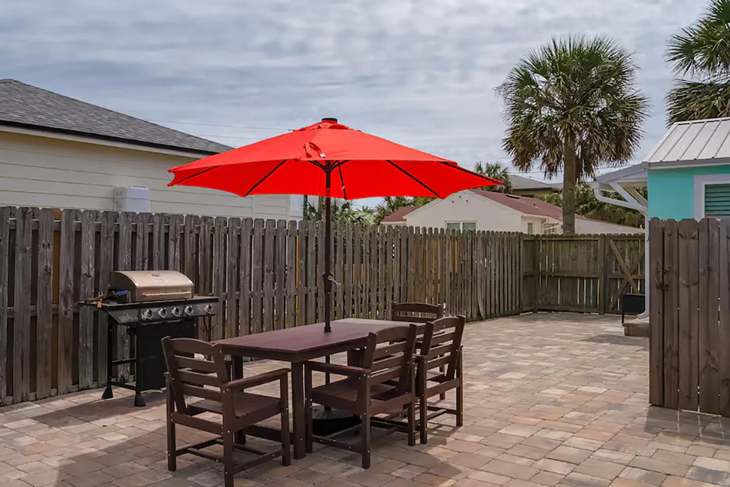 Outdoor patio with a paved stone floor, wooden dining table with chairs under a red umbrella, a gas grill, and a wooden privacy fence.