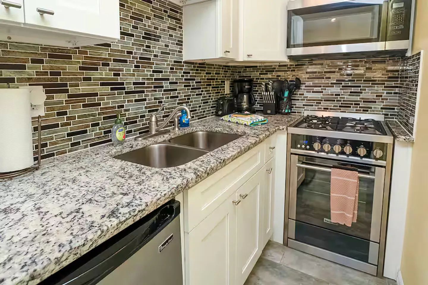 Kitchen corner with speckled granite countertops, double stainless steel sink, gas range, microwave, and multicolor glass mosaic tile backsplash.