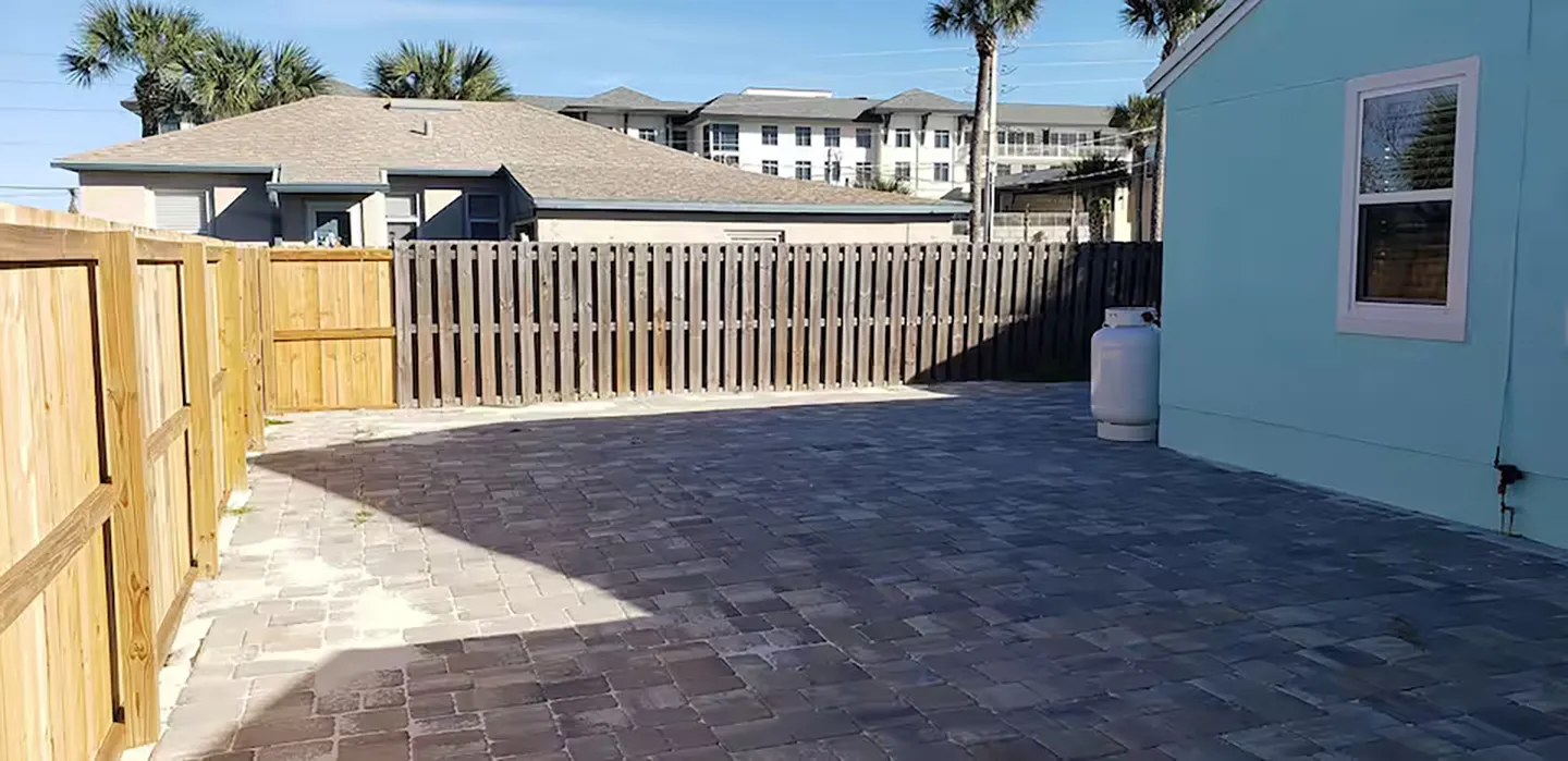 Fenced-in backyard patio paved with stone pavers, bordered by a wooden fence and the side of a light blue house.