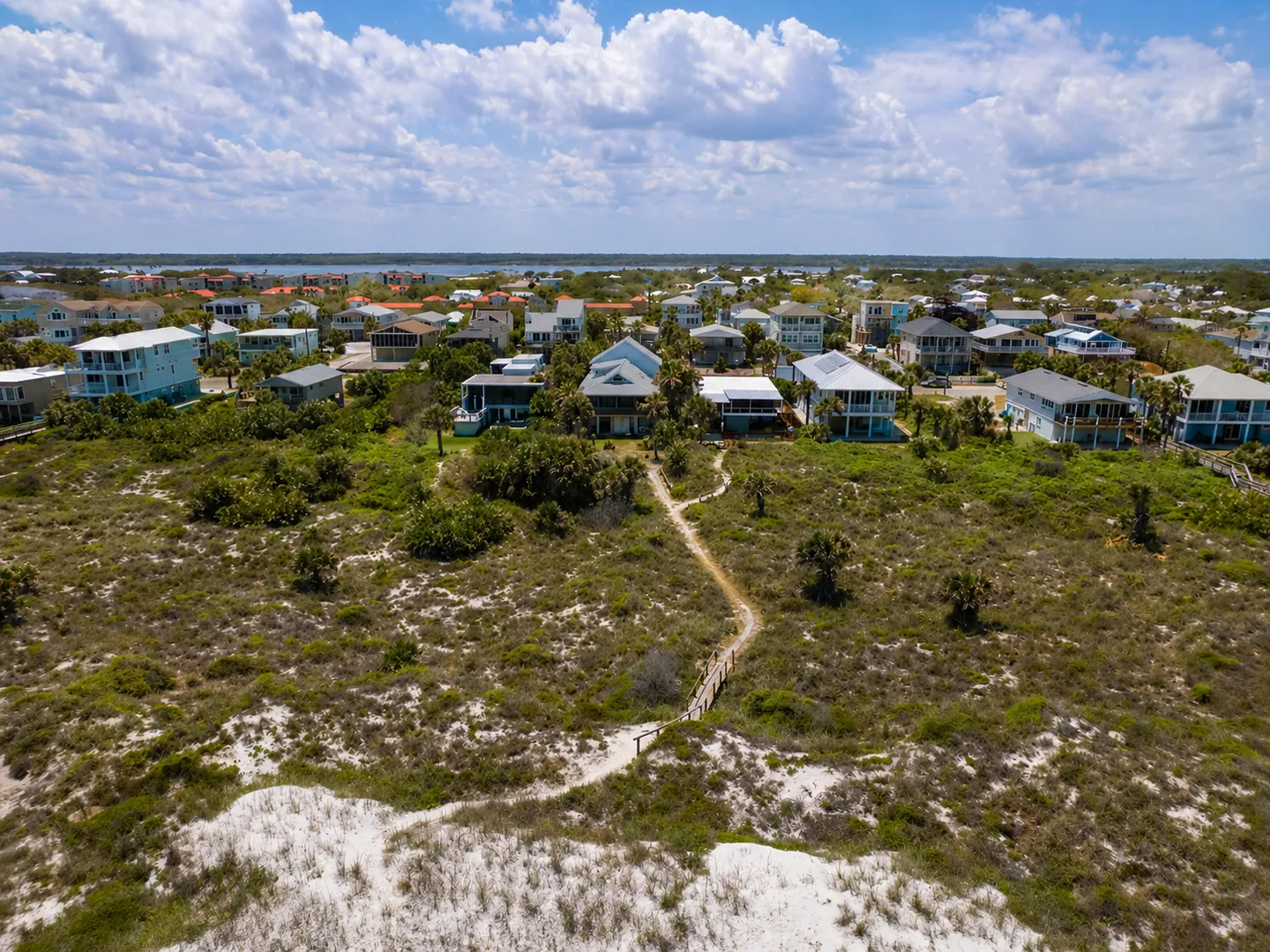 Aerial view of a coastal neighborhood with sand dunes, vegetation, and a wooden footpath leading to homes.